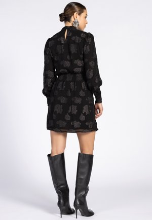 Zwarte jurk met lange mouwen, doorzichtige stof, metallic bloempatroon, aansluitende taille, keyhole achterkant, gecombineerd met zwart leren hoge hakken tot de knie.