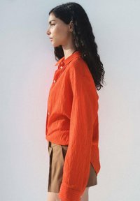 Orange texturerad långärmad skjorta med knäppt front, i kombination med bruna shorts. Synlig krage och kontrasterande sömmar.