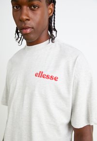 T-shirt en coton gris clair à manches courtes, avec un logo "ellesse" rouge sur le côté gauche de la poitrine, texture lisse et coupe décontractée.