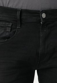 Prim-plan al secțiunii taliei unui jean negru din denim, cu buton și fermoar, arătând buzunarul frontal și bucla pentru curea pe un fundal gri.