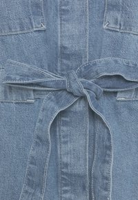 Tissu en denim bleu avec un nœud de ceinture noué à la taille et des coutures visibles sur de grandes poches avant et des coutures.