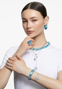 Tyk turkis smykkesæt bestående af en chunky halskæde, matchende armbånd, ringe og øreringe, vist på en models hænder og hals.