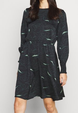 Žena v černých šatech po kolena s dlouhými rukávy a jemným vzorem zelených listů, s vypasovaným pasem a manžetovými detaily.