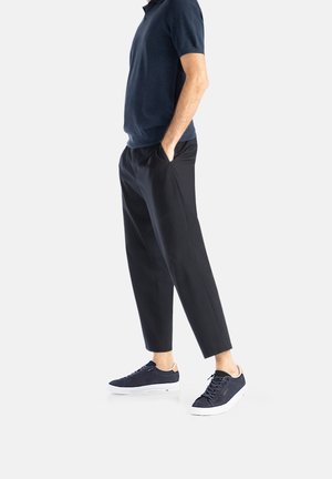 Polo shirt navy abbinato a pantaloni tapered neri e sneaker navy. Il materiale sembra morbido; le sneaker presentano suole bianche e dettagli beige.