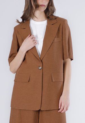 Mujer con una chaqueta marrón de manga corta con un solo botón sobre una camiseta blanca, y shorts marrones a juego.