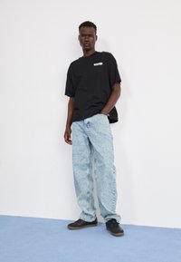 Črna oversize majica s belim logotipom, svetlo modri raztrgani široki jeans in temni športni čevlji. Enobarvna bela ozadje in modra tla.