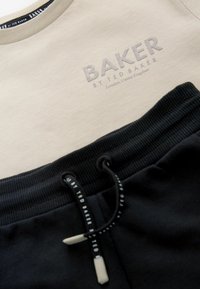 Šedé tričko s okrúhlym výstrihom a vykrojeným logom "BAKER" vyniká na béžovom materiáli nad čiernymi teplákmi s rebrovaným pásom a šnúrkovými detailmi.