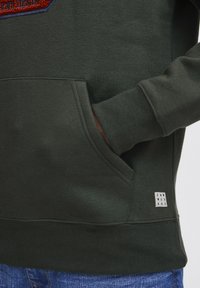 Green sweatshirt with kangaroo pocket, ribbed cuffs, and hem. Features a textured logo patch on the chest and a small brand label on the side.