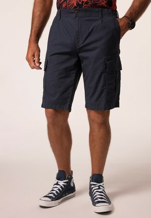 Man wearing dark blue cargo shorts and navy high-top sneakers, standing with one hand in pocket and one hand relaxed by side.