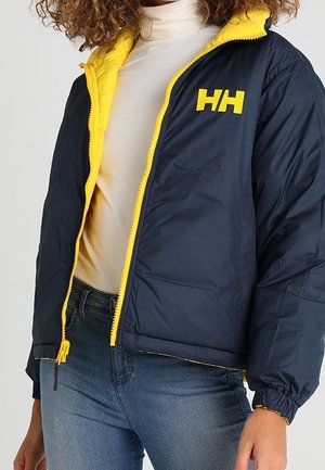 Femme portant une veste zippée bleu marine et jaune avec le logo HH, un col roulé blanc et un jean bleu, debout devant un fond uni.