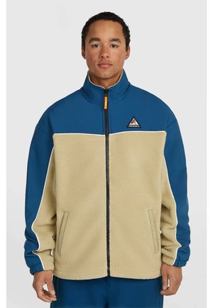 Jonge man draagt een blauw-beige fleecevest met rits, hoge kraag en logo op de borst tegen een grijze achtergrond.