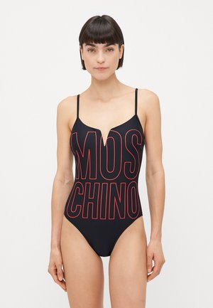 Naisso, kes kannab musta ühes tükis ujumistrikoo õhukeste rihmadega ning suurt punase äärisega "MOSCHINO" teksti ees, mille taust on lihtne.