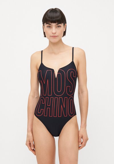 Kobieta w czarnym jednoczęściowym stroju kąpielowym z cienkimi ramiączkami i dużym, czerwonym napisem "MOSCHINO" na przodzie, na jednolitym tle.