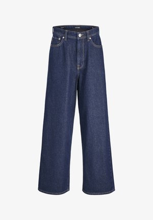 Personne portant une chemise blanche à manches courtes, un jean bleu à jambes larges, des baskets blanches et un foulard bleu clair à motifs autour du cou.