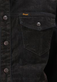 Črna denim jakna z srebrnimi gumbi, žepom na prsih s črto in oznako Wrangler v oranžni barvi.