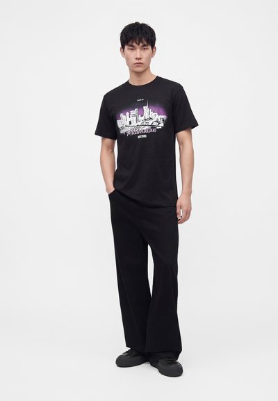 Jeune homme portant un t-shirt noir avec un graphisme de skyline urbain violet et blanc et un pantalon large noir sur fond blanc.