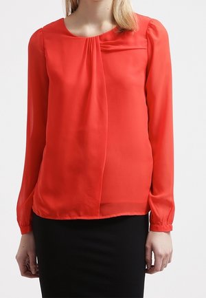 Frau trägt eine langärmelige, leuchtend rote Bluse mit Falten nahe dem Ausschnitt, kombiniert mit einem schwarzen Rock vor einem einfarbigen Hintergrund.