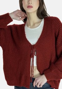 Rostfarbener Strickcardigan mit V-Ausschnitt, Bindebandverschluss und weitem Ärmeln. Texturiertes Muster ist überall sichtbar, getragen über einem weißen Oberteil.