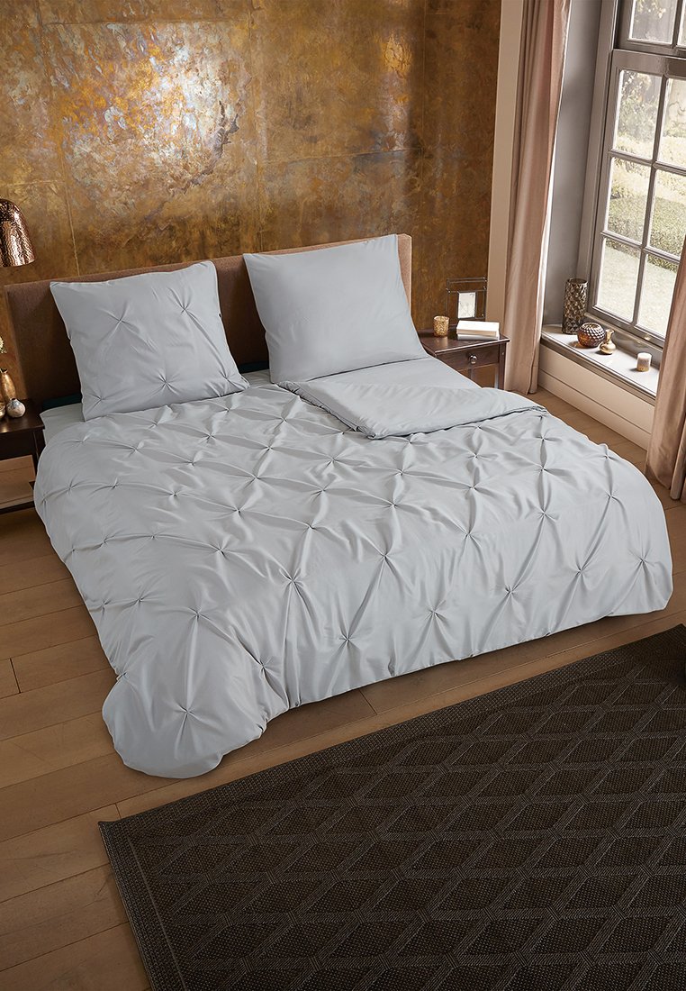 Ensemble de literie gris clair avec texture capitonnée, comprenant deux oreillers, une couette et un plaid décoratif sur un cadre de lit en bois, avec un tapis à motifs en dessous.