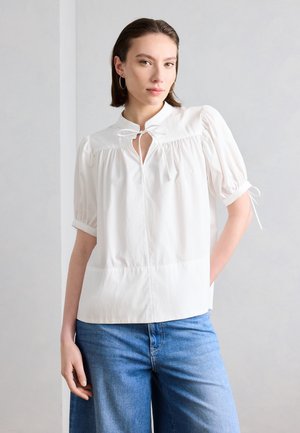 Vrouw met een witte blouse met puffmouwen en een strik aan de voorkant bij de kraag, en lichtblauwe broek met wijde pijpen, staand tegen een lichtgrijze muur.