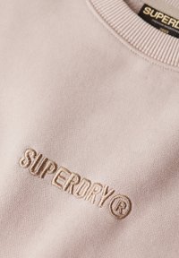 Lyserød sweatshirt med ribbet halsudskæring og broderet "SUPERDRY" logo i beige. Blødt stof med en glat tekstur.