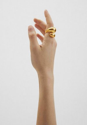 Hand mit erhobenen Fingern, die einen offenen Goldring mit zwei glatten, abgerundeten Enden vor einem schlichten hellen Hintergrund trägt.