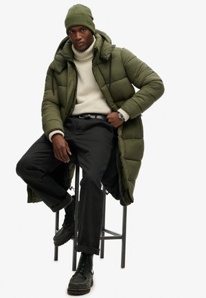 Manteau rembourré vert olive avec un col haut, pull beige clair, pantalon noir et bottes noires. Modèle assis sur un tabouret haut.