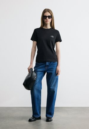 Sieviete melnā Calvin Klein t-kreklā, zilās džinsās, melnās loafer kurpēs, saulesbrillēs, turot melnu ādas somu vienkāršā fonā.