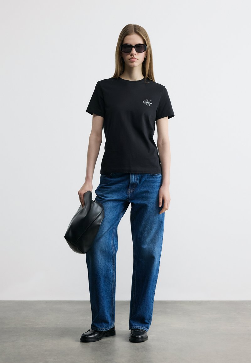 Γυναίκα που φοράει μαύρο μπλουζάκι Calvin Klein, μπλε τζιν, μαύρα μοκασίνια, γυαλιά ηλίου, κρατώντας μαύρη δερμάτινη τσάντα σε απλό φόντο.