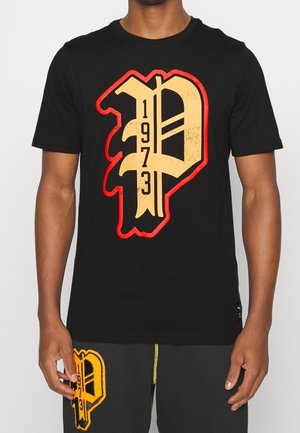 Hombre con camiseta negra con gran contorno amarillo y rojo de "P" y números 1973, logo a juego en pantalones negros.
