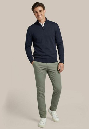 Glimlachende man met een marineblauwe trui met halve rits, lichtgroene broek en witte sneakers, staand tegen een effen beige achtergrond.