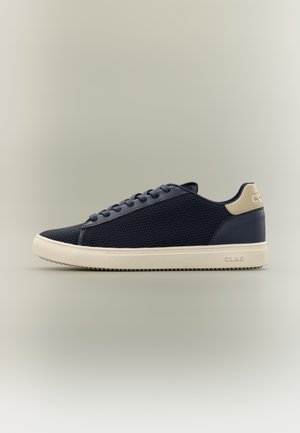Zapatilla baja azul marino con suela y lengüeta trasera beige, parte superior de malla y diseño con cordones, mostrada de perfil sobre un fondo neutro.