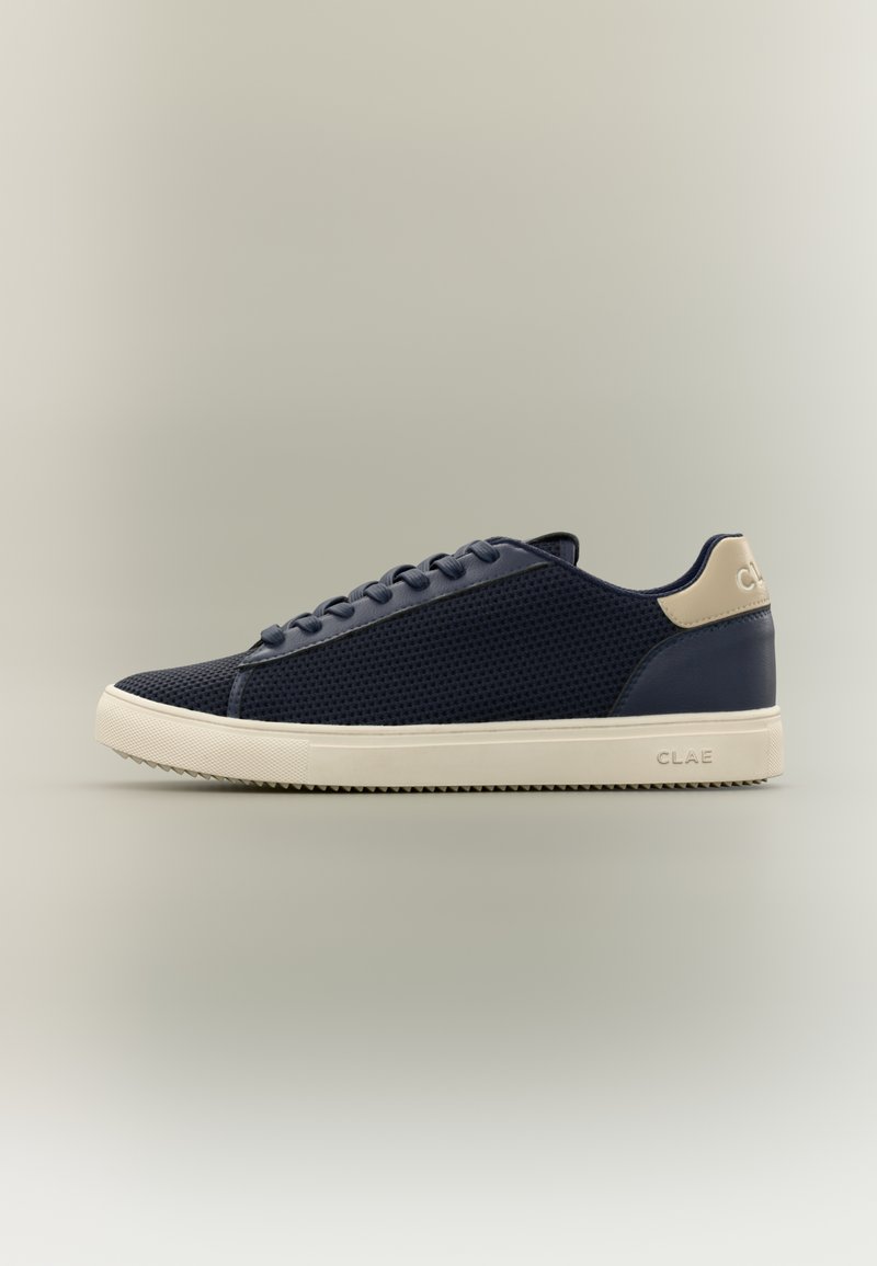 Παπούτσι τύπου sneaker χαμηλού προφίλ σε ναυτικό μπλε χρώμα με μπεζ σόλα και ταμπελάκι στη φτέρνα, πλέγμα στο πάνω μέρος και σχέδιο με κορδόνια, εμφανιζόμενο σε πλαινή προβολή σε ουδέτερο φόντο.