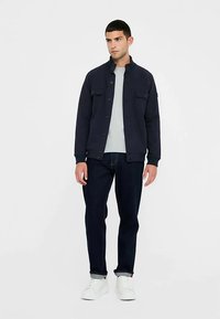 Donkerblauwe jas met kraag en voorzakken, gecombineerd met een lichtgrijs shirt en donkere spijkerbroek. Witte sneakers maken de outfit compleet.