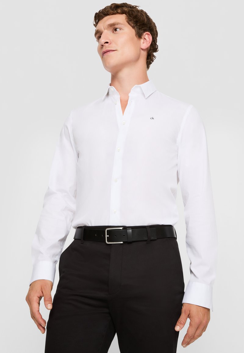 Weißes Hemd mit Knöpfen aus Baumwollstoff, langärmlig, mit spitzem Kragen. Kleines Logo auf der Brust, kombiniert mit einem schwarzen Gürtel und einer Hose.