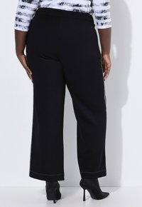 Kobieta ubrana w czarne spodnie o szerokich nogawkach i czarne szpilki, stojąca na tle białej ściany, w czarno-białej bluzce z wzorem.