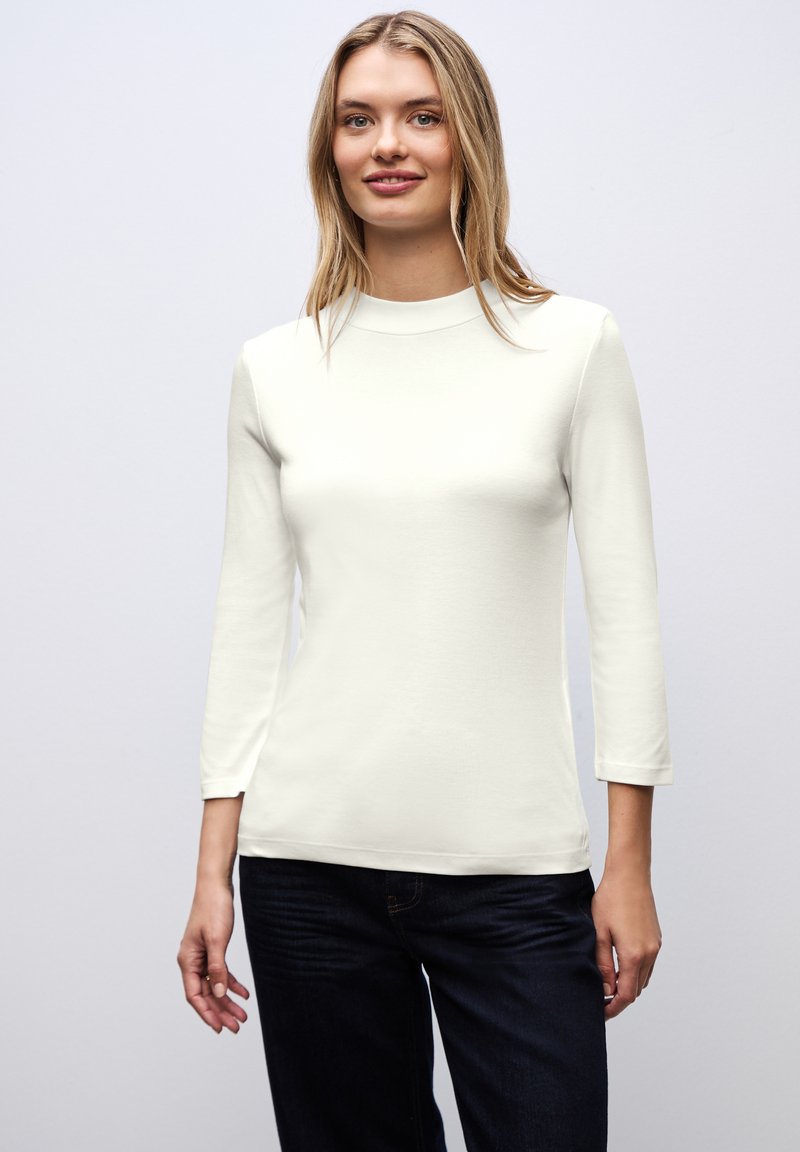 Camiseta blanca de manga larga con escote alto. Material suave y ajustado, con mangas tres cuartos, combinado con jeans de color azul oscuro.