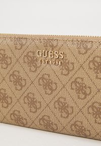 Brown textured wallet with diamond pattern, featuring gold "GUESS" logo and "EST 1981" detail, secured with a gold zipper.