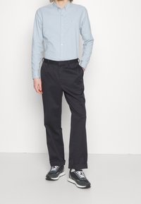 Ljusblå skjorta med knappar, mörkgrå byxor och svarta sneakers. Skjortan har en liten logotyp på bröstet, slätt tyg och en avslappnad passform.