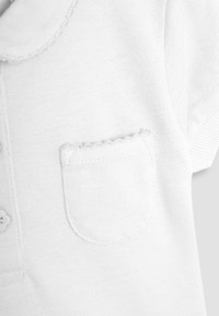 Fehér, texturált rövid ujjú pólóing, kisméretű zsebbel a mellrészen, és hullámos szegéllyel a galléron és a zseb felső részén.