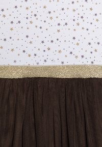 Brun tyllkjol med ett plisserat mönster. Översta delen har ett vitt tyg dekorerat med små grå stjärnor och gyllene gnistror, kantat med metallisk guldfinish.