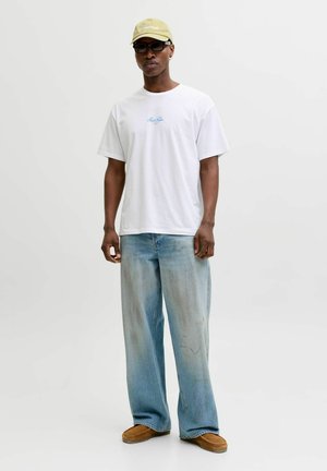 Muškarac s bež kapom, crnim sunčanim naočalama, bijelom majicom s plavim tekstom, širokim svijetloplavim trapericama i smeđim mokasinama stoji nasuprot jednobojnoj pozadini.