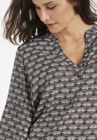 Blusa a fantasia nera con disegni a ventaglio bianchi e ruggine. Presenta un collo a V e un tessuto morbido e strutturato. Vestibilità larga con maniche fluide.