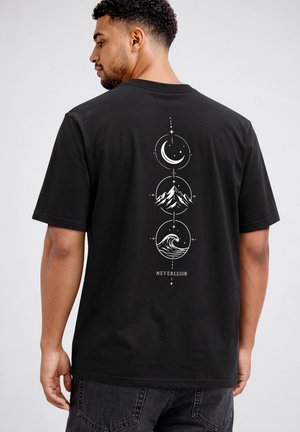 Mann trägt schwarzes T-Shirt mit vertikalem weißen Design eines Halbmonds, Bergs und Ozeanwelle sowie dem Wort „NEVERLESS“ darunter.