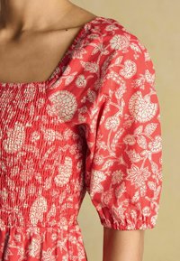 Rotes Blumenkleid mit quadratischem Ausschnitt, kurzen Puffärmeln und gerafftem Oberteil mit weißen Blumenmustern auf einem strukturierten Stoff.