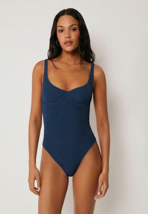 Frau mit langen dunklen Haaren, die einen strukturierten marineblauen Einteiler trägt und vor einem schlichten hellen Wandhintergrund steht.