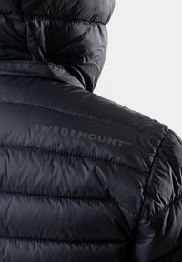 Sort dunjakke med hætte, der har quiltet tekstur og glat stof. Mærkenavnet "SWEDEMOUNT" er præget på skulderen.