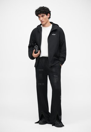 Schwarze Zip-Jacke mit Kapuze, darunter ein weißes T-Shirt, schwarze Hose mit grafischen Prints und schwarze Sneaker. Glatte Textur insgesamt.