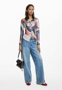 Long-sleeve, multicolored, button-up shirt with abstract pattern, paired with wide-leg blue jeans and red slip-on shoes. Black handbag with chain strap.