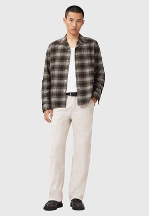 Young man wearing brown and gray plaid button-up shirt, white tank top, beige pants, black belt, and black shoes standing with one hand in pocket.